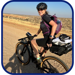 This photograph show Franci in action riding her bicycle, looking back at the photographer and smiling. She is wearing a black athletic outfit, sunglasses, and safety gear. She has light skin and brown hair.