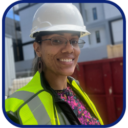 Photo of Janei looking at the camera and smiling. Janei has brown skin, wears glasses, and has her hair pulled back. She is wearing a pink floral top, under a safety vest and a hard hat due to her career in development.